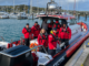 Coastguard Mana volunteers in red rescue gear stand aboard Te Awarua Rescue at Mana Marina, with yachts and moored boats in the background.