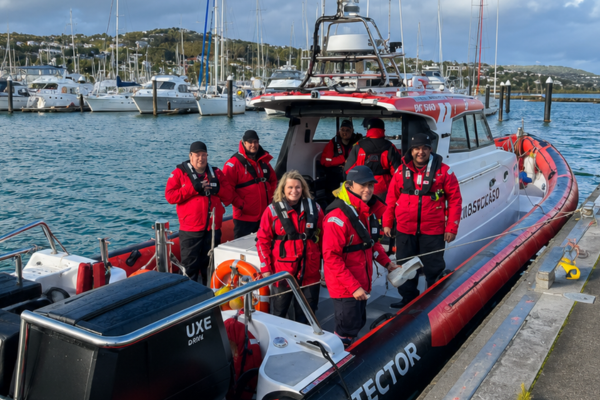 Coastguard Mana volunteers in red rescue gear stand aboard Te Awarua Rescue at Mana Marina, with yachts and moored boats in the background.