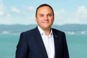 Former Porirua mayor Nick Leggett wearing a navy suit and white shirt, standing in front of a blurred waterfront backdrop.