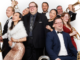 Porirua City Brass performers pose in formal and cabaret-style clothing, smiling and holding brass instruments.