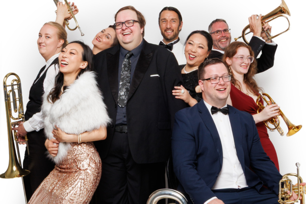 Porirua City Brass performers pose in formal and cabaret-style clothing, smiling and holding brass instruments.