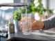A hand holds a clear glass under a chrome kitchen faucet as water fills it.