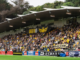 Wellington Phoenix fans fill the grandstand at Porirua Park during an A-League Women match, waving yellow-and-black flags and banners ahead of the club’s semi-final showdown with Brisbane Roar.
