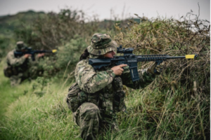 Two New Zealand Army Reserve soldiers in camouflage move through long grass and scrub during a field training exercise.