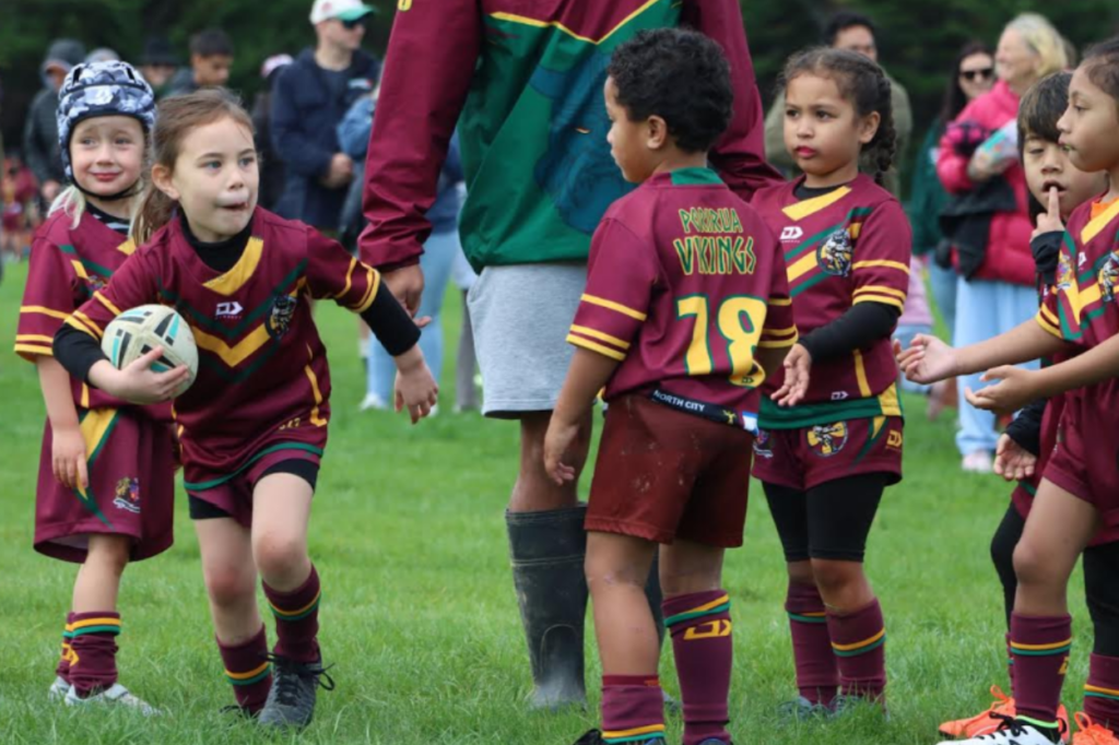 Young Porirua Vikings junior rugby league players in maroon and green uniforms take part in a game on a grass field, with one child running forward carrying the ball.