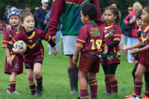 Young Porirua Vikings junior rugby league players in maroon and green uniforms take part in a game on a grass field, with one child running forward carrying the ball.