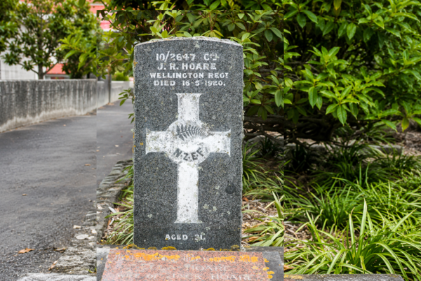 A grey stone war grave headstone for Corporal J. R. Hoare of the Wellington Regiment, partly weathered, beside a path and greenery at Porirua Cemetery.