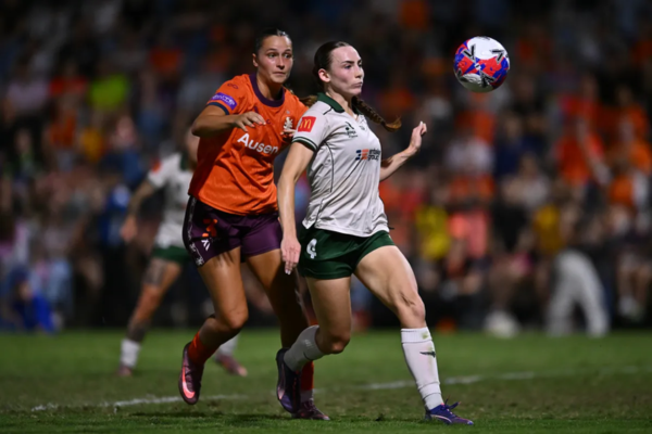 Wellington Phoenix defender tracks the ball in mid-air while a Brisbane Roar player pressures from behind during a semi-final match.