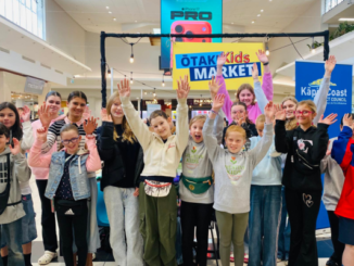 A group of smiling children raise their hands in front of an Ōtaki Kids Market stall at Coastlands Shopping Centre.