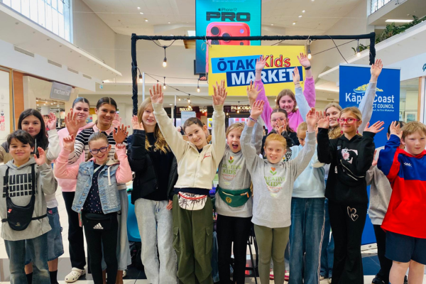 A group of smiling children raise their hands in front of an Ōtaki Kids Market stall at Coastlands Shopping Centre.