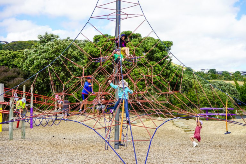Children climb and play on a large rope climbing structure at a playground, surrounded by trees and open space.