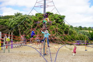 Children climb and play on a large rope climbing structure at a playground, surrounded by trees and open space.