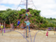 Children climb and play on a large rope climbing structure at a playground, surrounded by trees and open space.