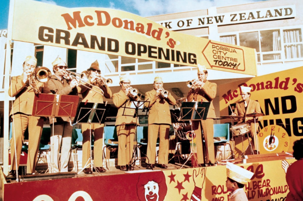 A brass band performs on a stage during the grand opening of New Zealand’s first McDonald’s restaurant in Porirua on June 7, 1976. Large "Grand Opening" signs and McDonald’s branding surround the stage as crowds gather to celebrate.