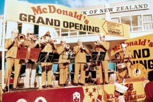 A brass band performs on a stage during the grand opening of New Zealand’s first McDonald’s restaurant in Porirua on June 7, 1976. Large "Grand Opening" signs and McDonald’s branding surround the stage as crowds gather to celebrate.