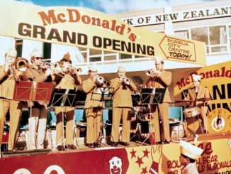 A brass band performs on a stage during the grand opening of New Zealand’s first McDonald’s restaurant in Porirua on June 7, 1976. Large "Grand Opening" signs and McDonald’s branding surround the stage as crowds gather to celebrate.
