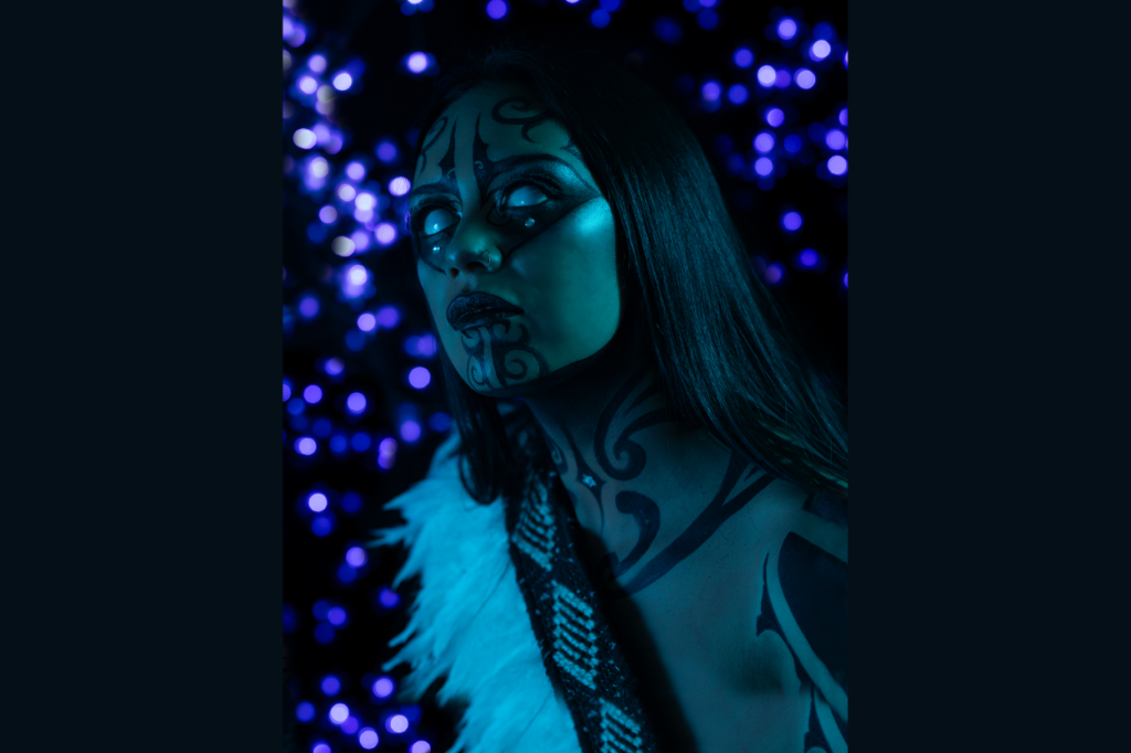 Portrait of a person painted with black Matariki-inspired face and body designs, lit in blue, with purple lights behind them.