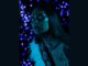 Portrait of a person painted with black Matariki-inspired face and body designs, lit in blue, with purple lights behind them.
