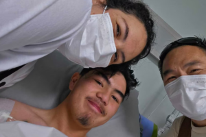 Ashen Phaipanya smiling in a hospital bed in Bali with his parents, Sheena and Ants, beside him.