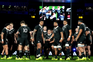 All Blacks players gather on the field during a rugby match, with the stadium screen visible in the background.