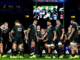 All Blacks players gather on the field during a rugby match, with the stadium screen visible in the background.