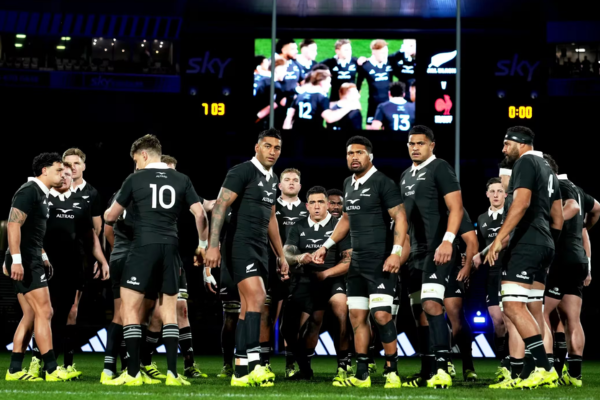 All Blacks players gather on the field during a rugby match, with the stadium screen visible in the background.