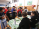Young people wearing aprons talk with seated adults during a community activity in a Porirua hall.