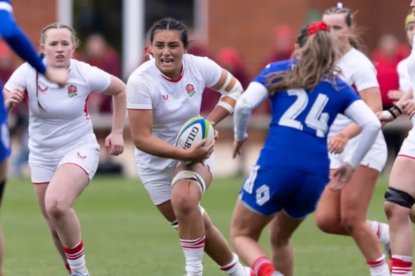An England women’s age-group rugby player carries the ball towards a defender in blue during a match.