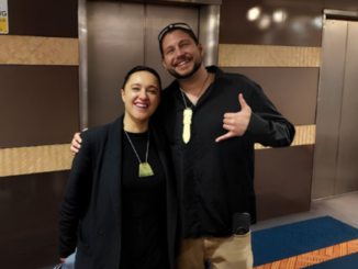 Two people smile with their arms around each other, both wearing pounamu pendants, in a building foyer.