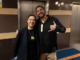 Two people smile with their arms around each other, both wearing pounamu pendants, in a building foyer.