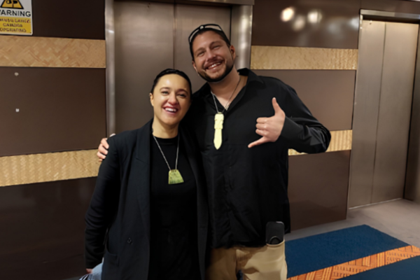 Two people smile with their arms around each other, both wearing pounamu pendants, in a building foyer.