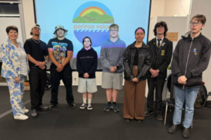 Group photo of Porirua students and an adult standing in front of a projected Porirua Voices Youth Podcast logo at the podcast launch event.