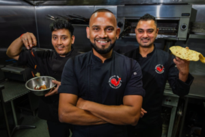 Three Sushil’s Musclechef Café team members in a commercial kitchen, with one holding a pan of food and another holding flatbread.
