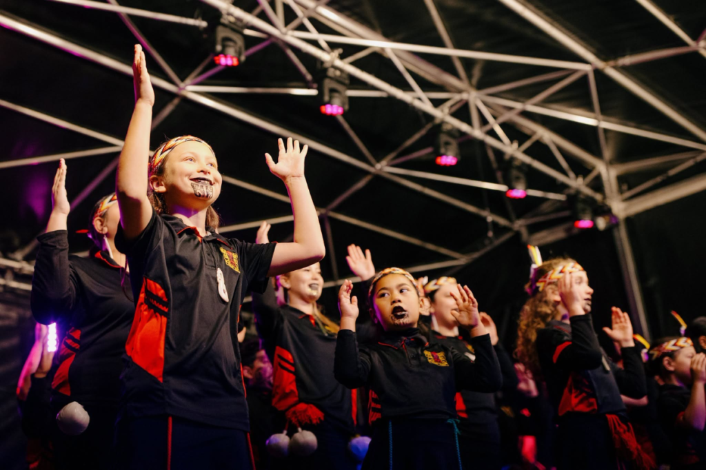 Young performers from Ngā Taitāmariki ō Ngāti Pōneke Young Māori Club perform on stage with raised hands.
