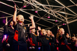 Young performers from Ngā Taitāmariki ō Ngāti Pōneke Young Māori Club perform on stage with raised hands.