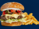 A cheeseburger topped with lettuce, tomato and onion, served with seasoned fries against a dark blue background.