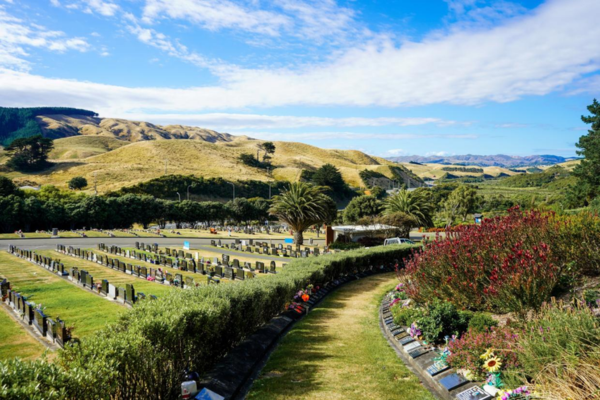 Rows of headstones and memorial plots at a Porirua cemetery, with landscaped gardens, palm trees and hills in the background.