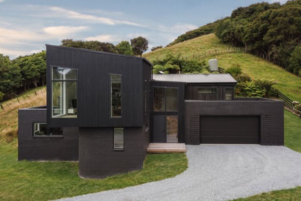 Deviation House in Pāuatahanui, with dark timber cladding, brick walls and a double garage set against a rural hillside.