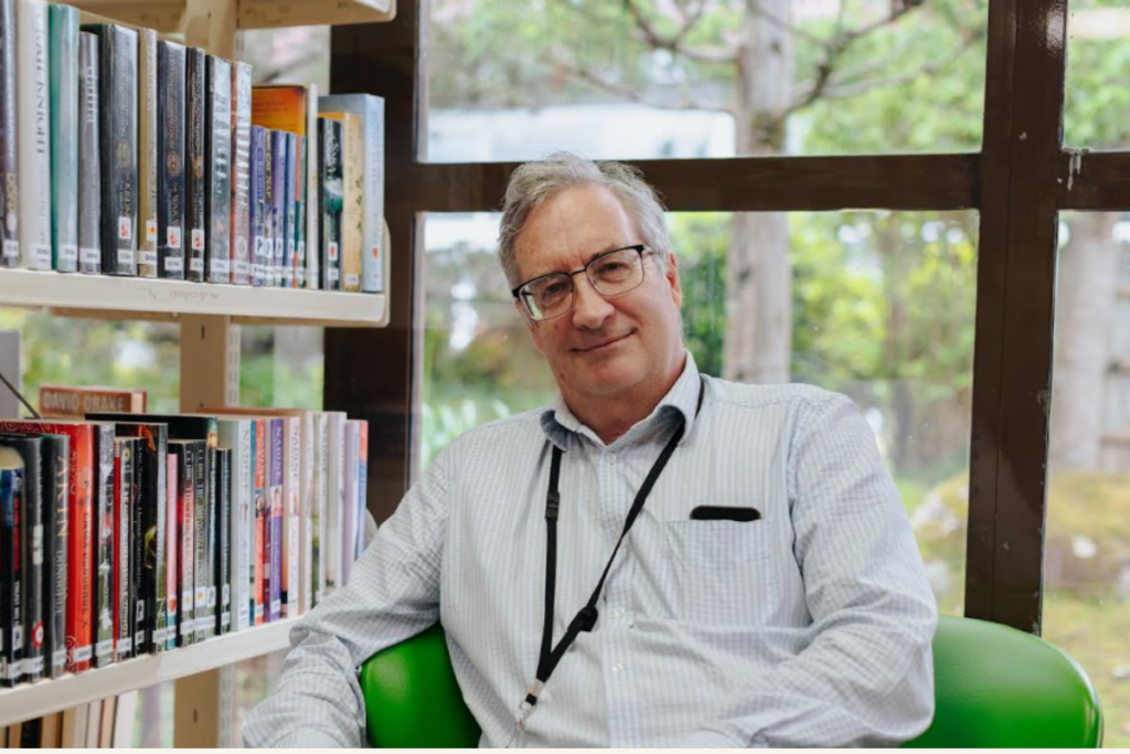 Porirua Libraries Manager Brian Anderson sitting inside a library surrounded by books.
