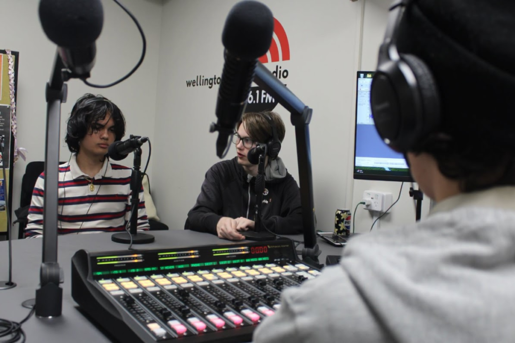 Three young people wearing headphones sit around microphones in a radio studio, with a mixing desk in the foreground.