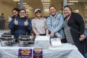 Five women smiling behind a food stall with trays of island food at a Pacific Night Market in Cannons Creek.