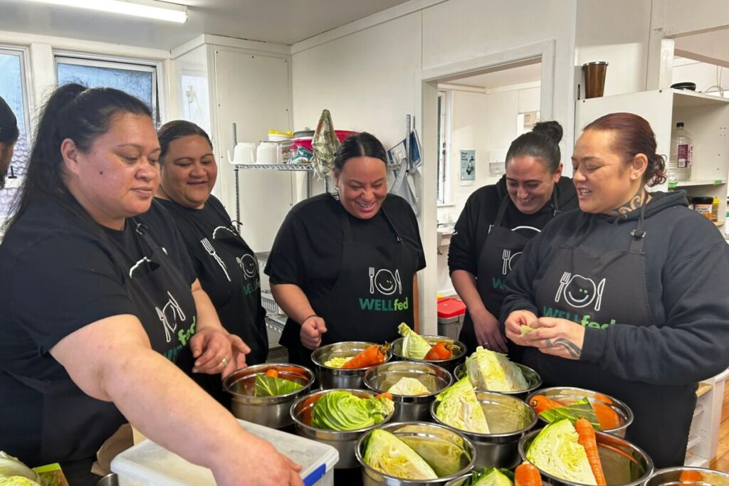 Five adults wearing WELLfed aprons prepare vegetables together in a kitchen.