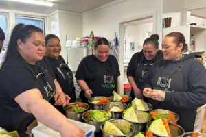 Five adults wearing WELLfed aprons prepare vegetables together in a kitchen.