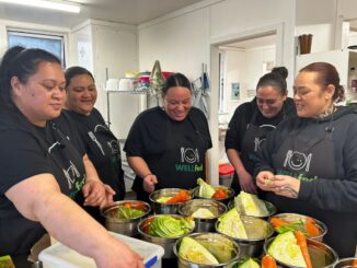 Five adults wearing WELLfed aprons prepare vegetables together in a kitchen.